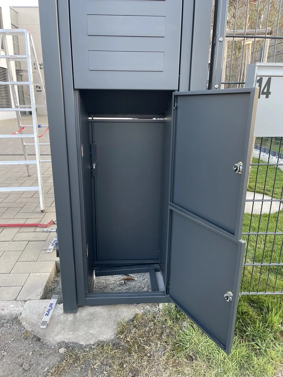 Offene graue Paketbox aus pulverbeschichtetem Stahl neben Zaun mit zweiflügeliger Tür