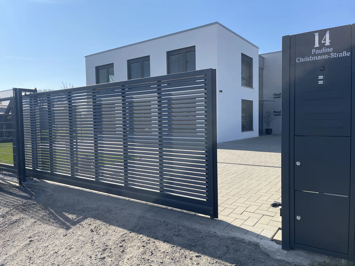 Modernes Schiebetor aus pulverbeschichtetem Metall mit Lamellen vor Einfahrt eines weißen Wohnhauses