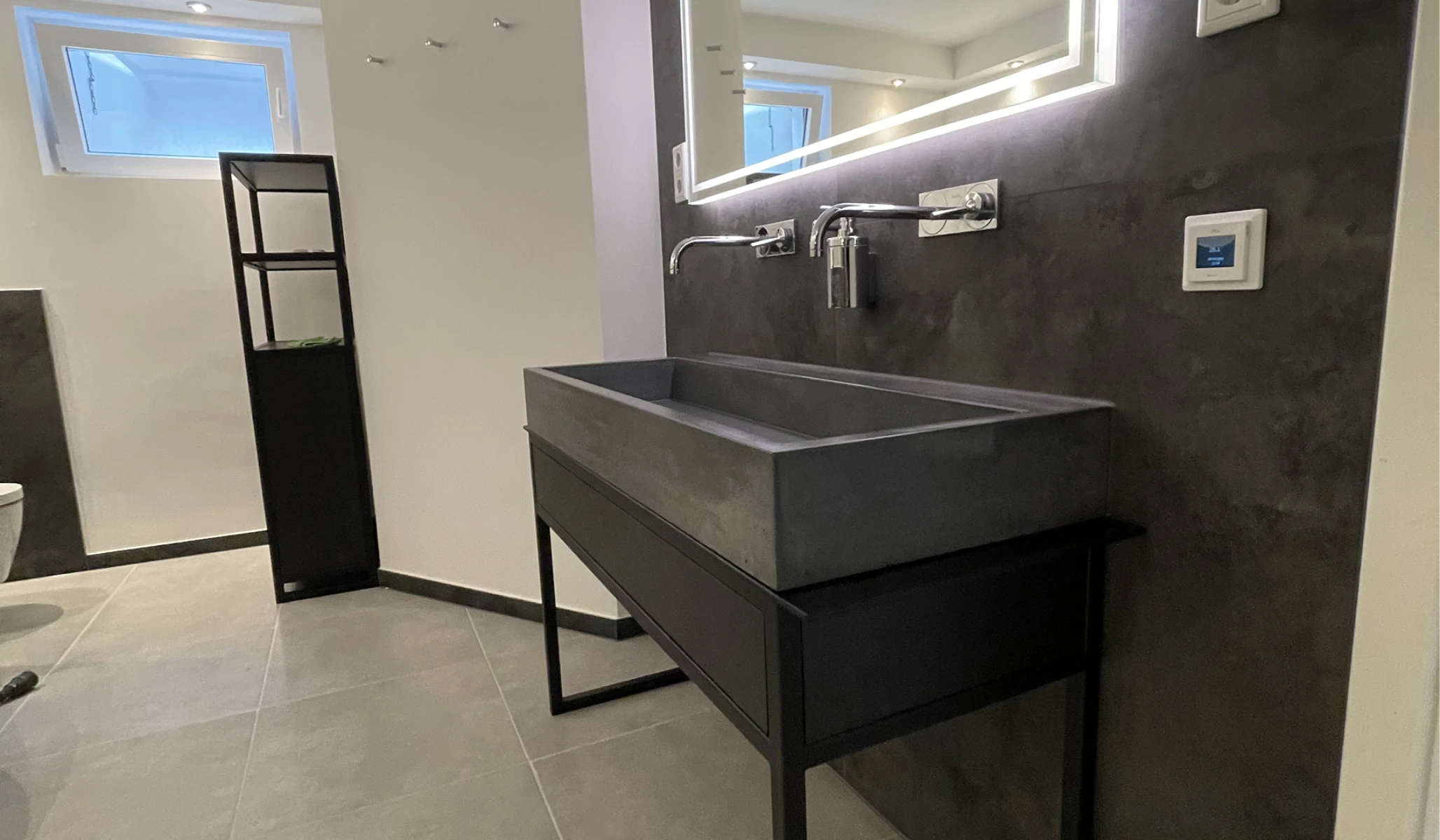 Modernes Bad mit rechteckigem Betonwaschbecken auf schwarzem Metallgestell, Wandarmaturen und Spiegel mit LED-Beleuchtung